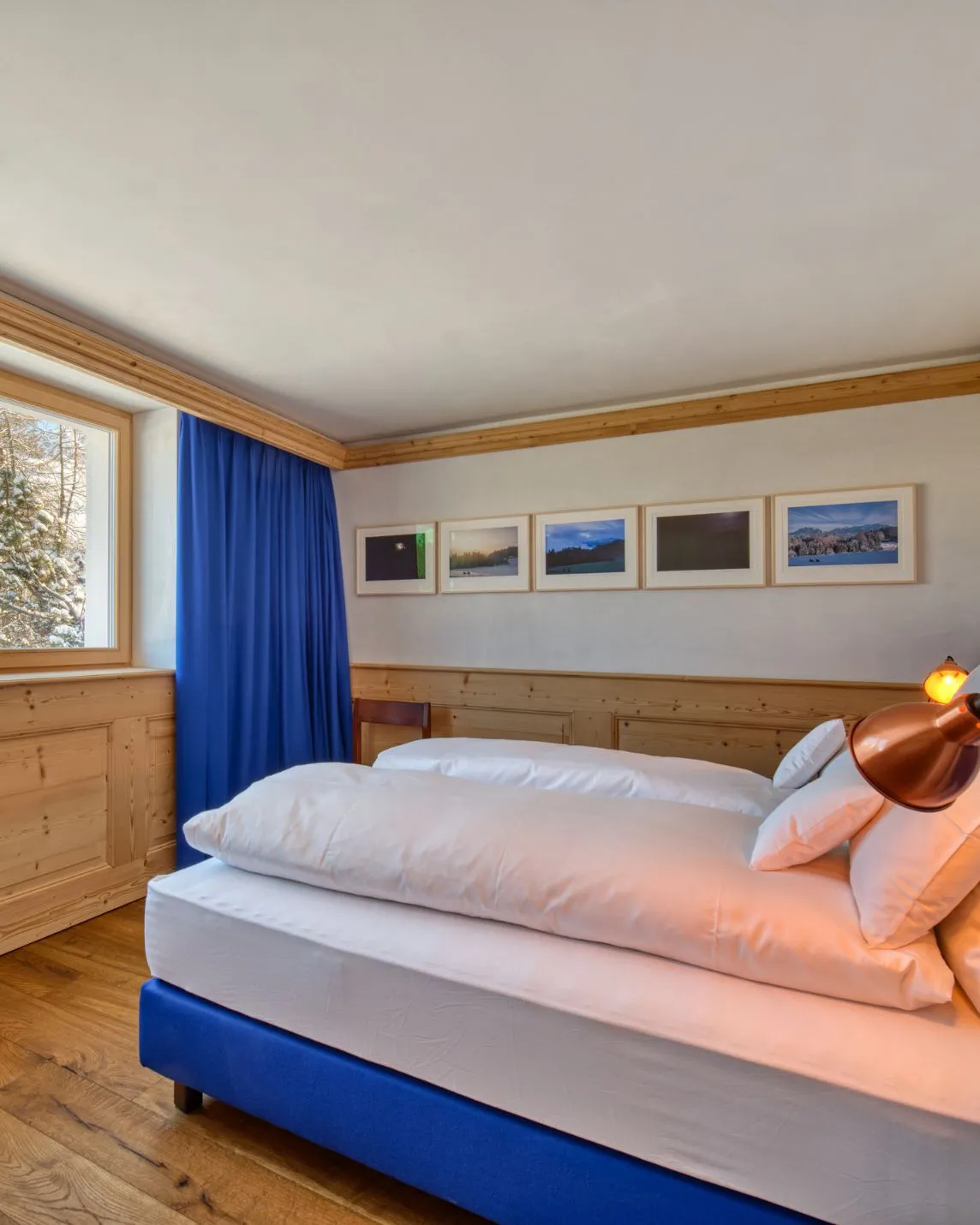 A cosy bedroom with a white duvet on a blue bed frame, blue curtains, wood panelling, five framed landscape photographs on the wall and a copper reading lamp
