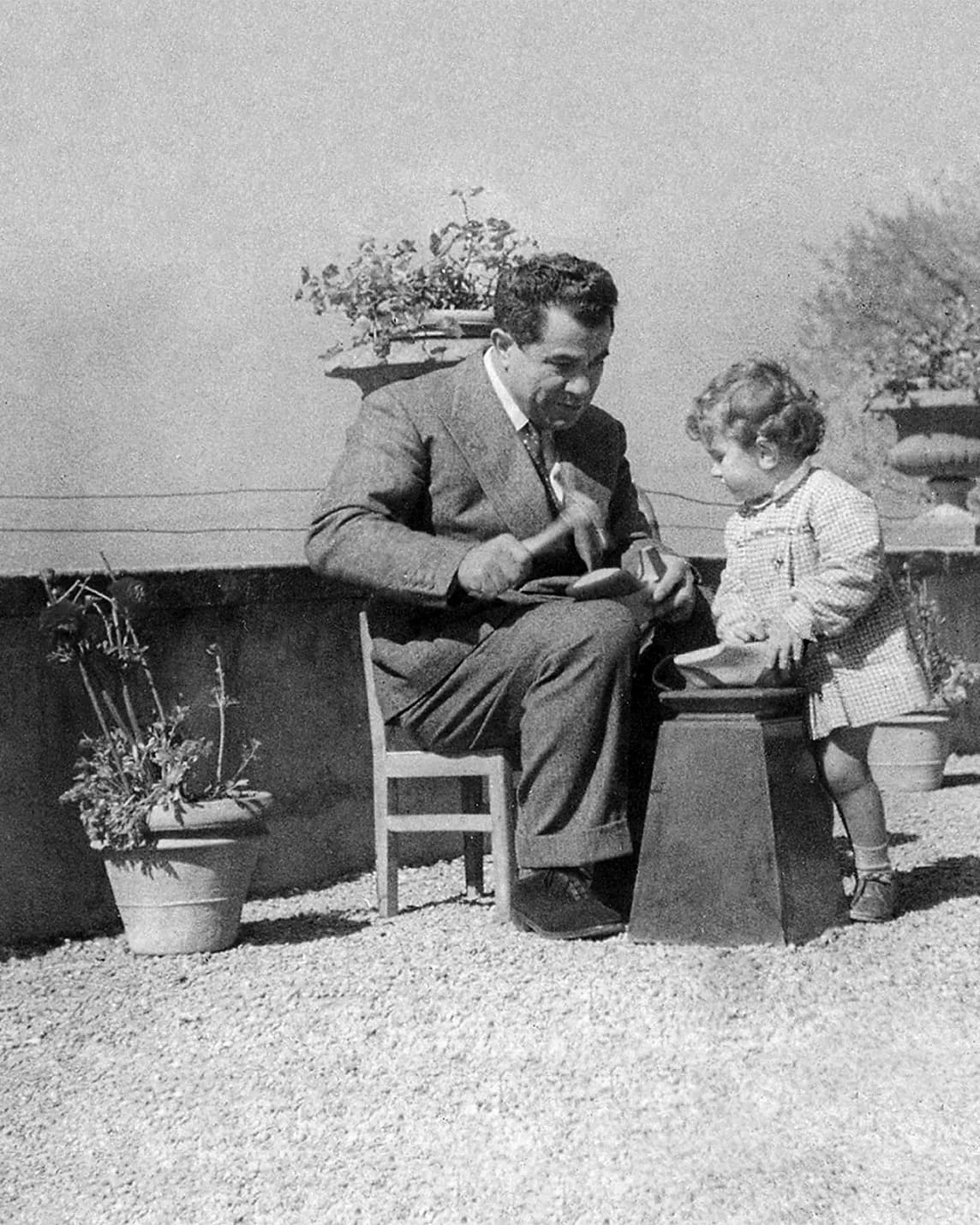 An adult man in a suit sits on a chair on a gravel terrace, reading with a young girl standing beside a pedestal; potted plants surround them.