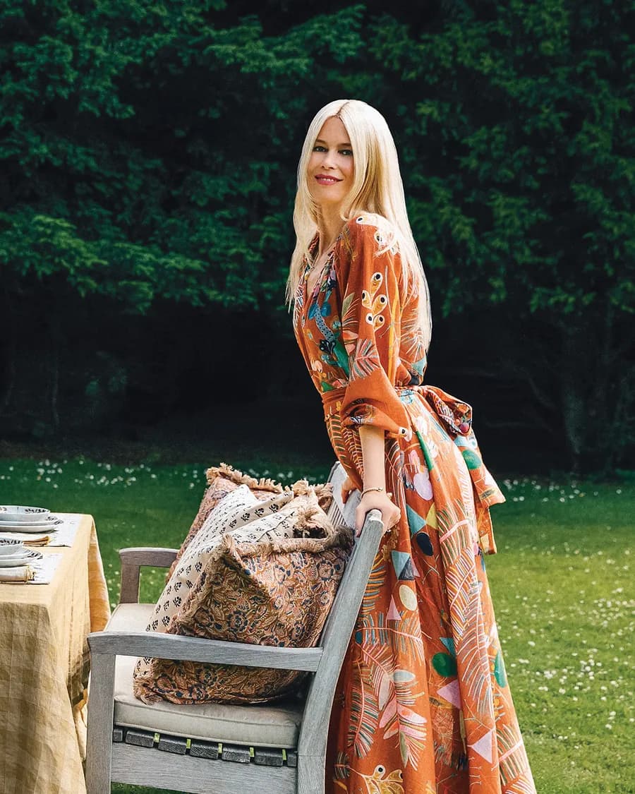 Blonde woman in a vibrant orange patterned dress stands beside a chair with decorative cushions in a sunny garden.