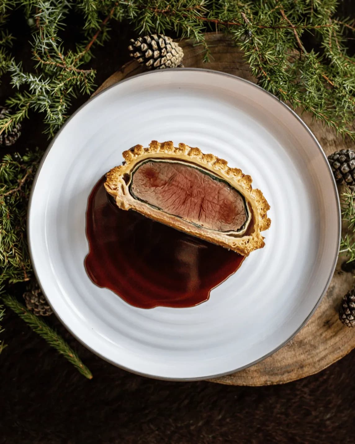 Beef Wellington slice with pink center on a white plate, with red wine sauce, set on a rustic board among evergreen branches and pinecones.