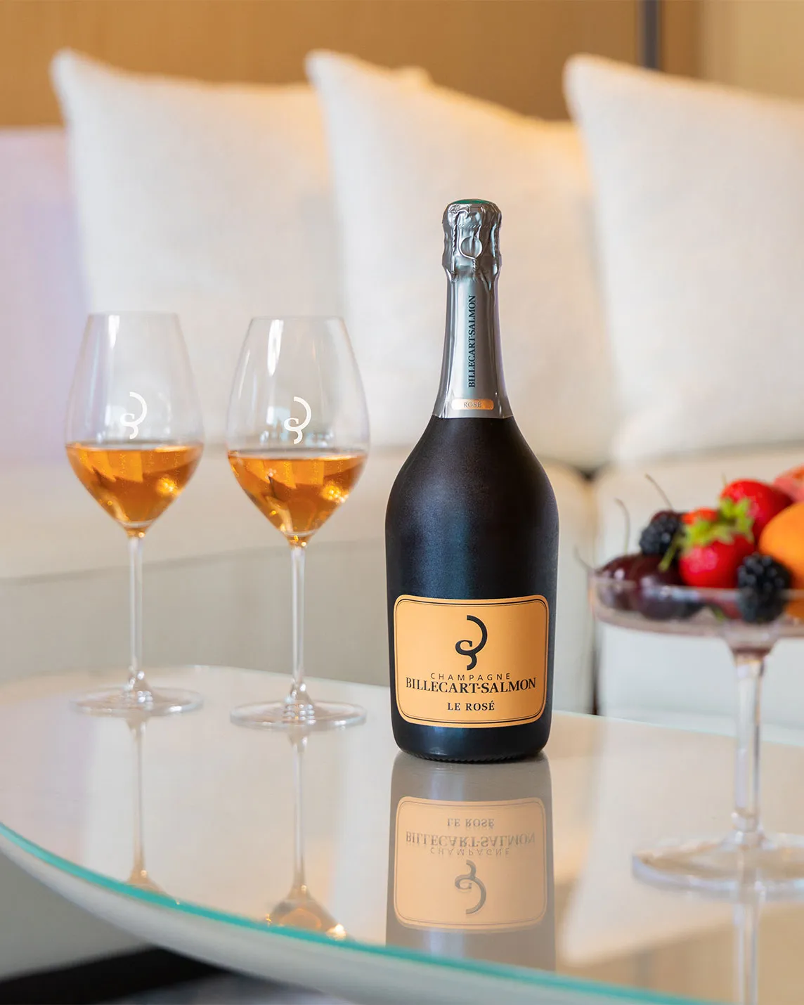 A bottle of Billecart-Salmon Le Rosé Champagne on a glass table with two filled glasses and a fruit bowl, soft cushions in the background.