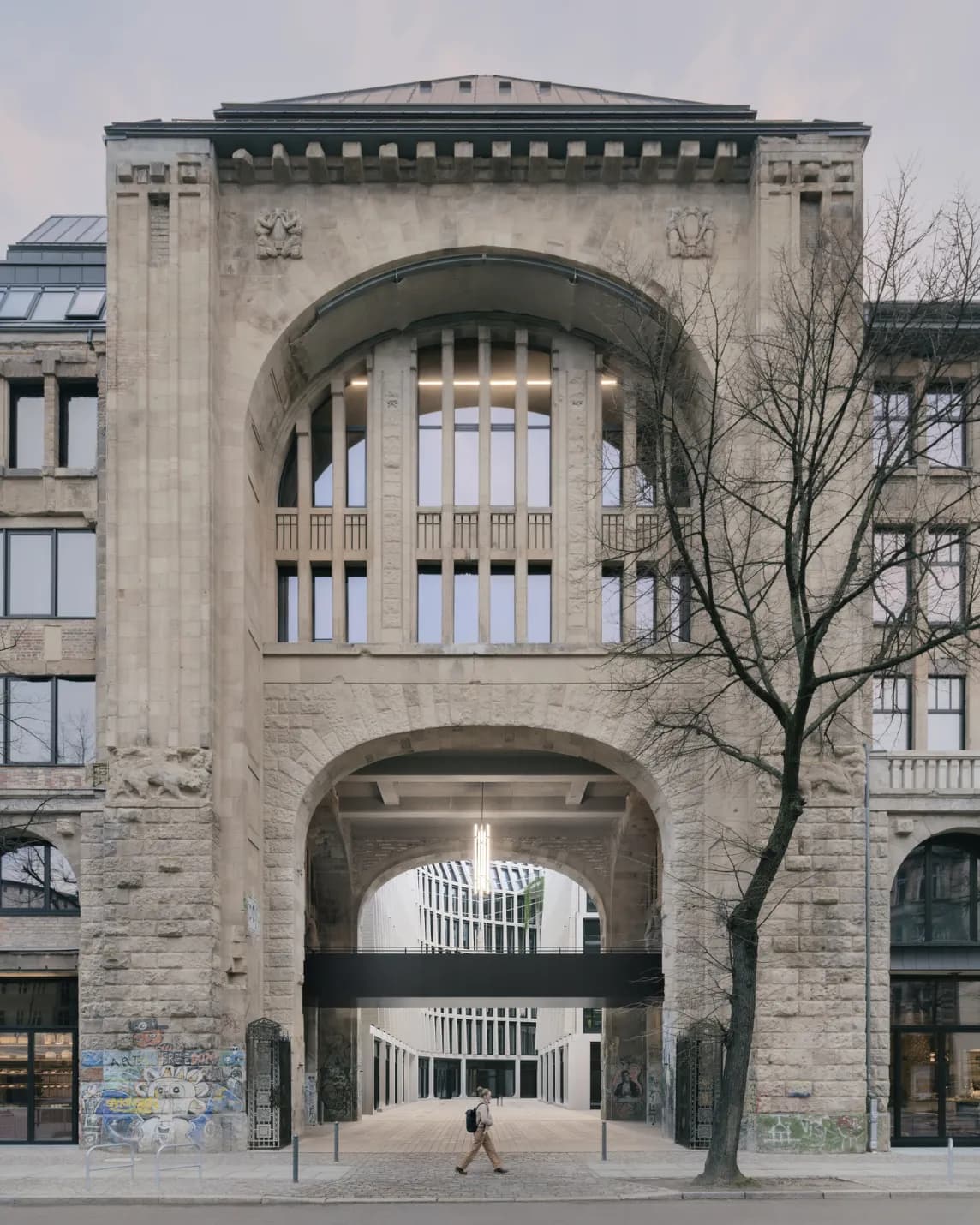 A grand stone archway with tall windows above frames a passage to a modern courtyard; a leafless tree stands to the right as a person walks by.