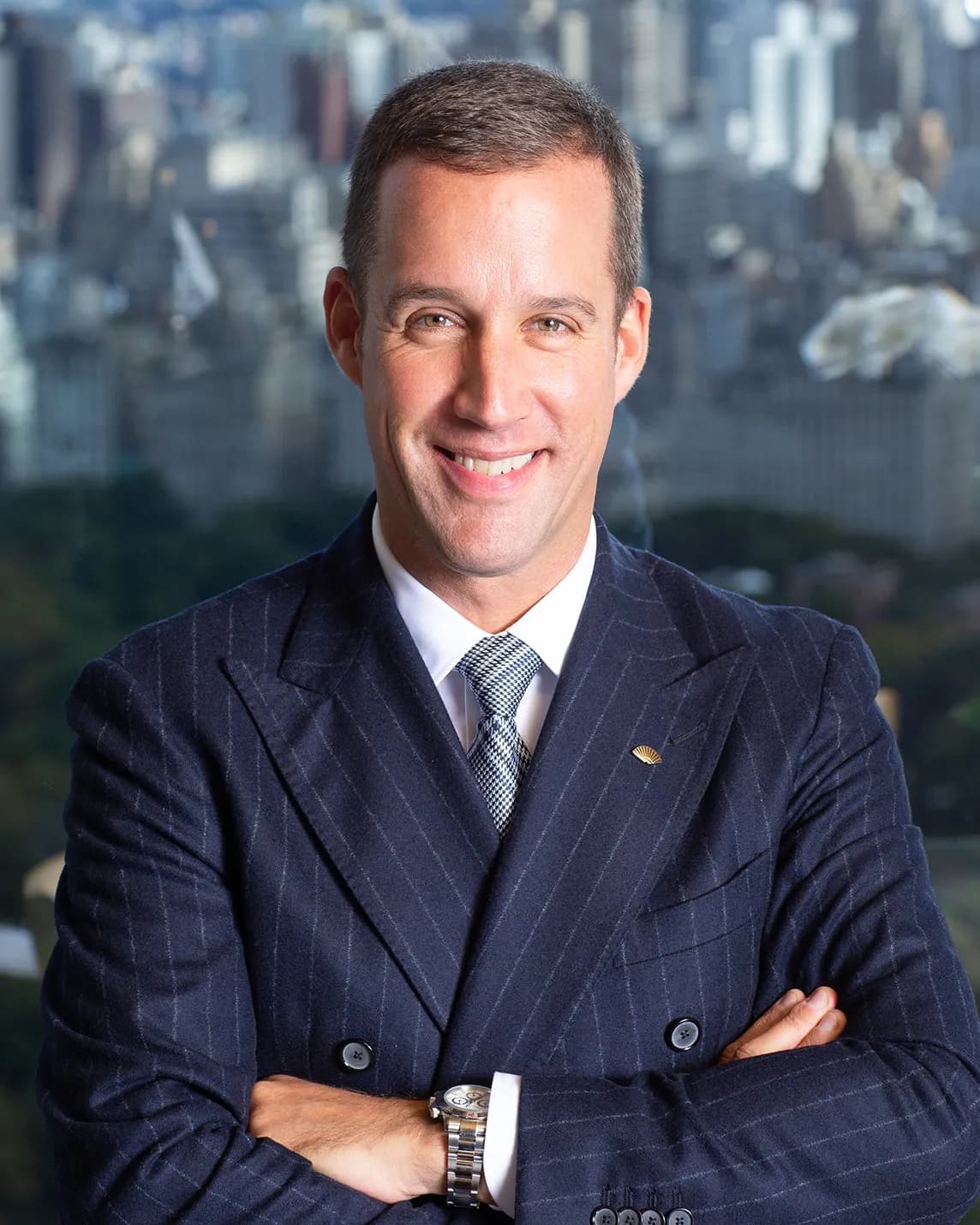 Smiling man in a dark pinstripe suit with a patterned tie, arms crossed, against a city skyline backdrop.