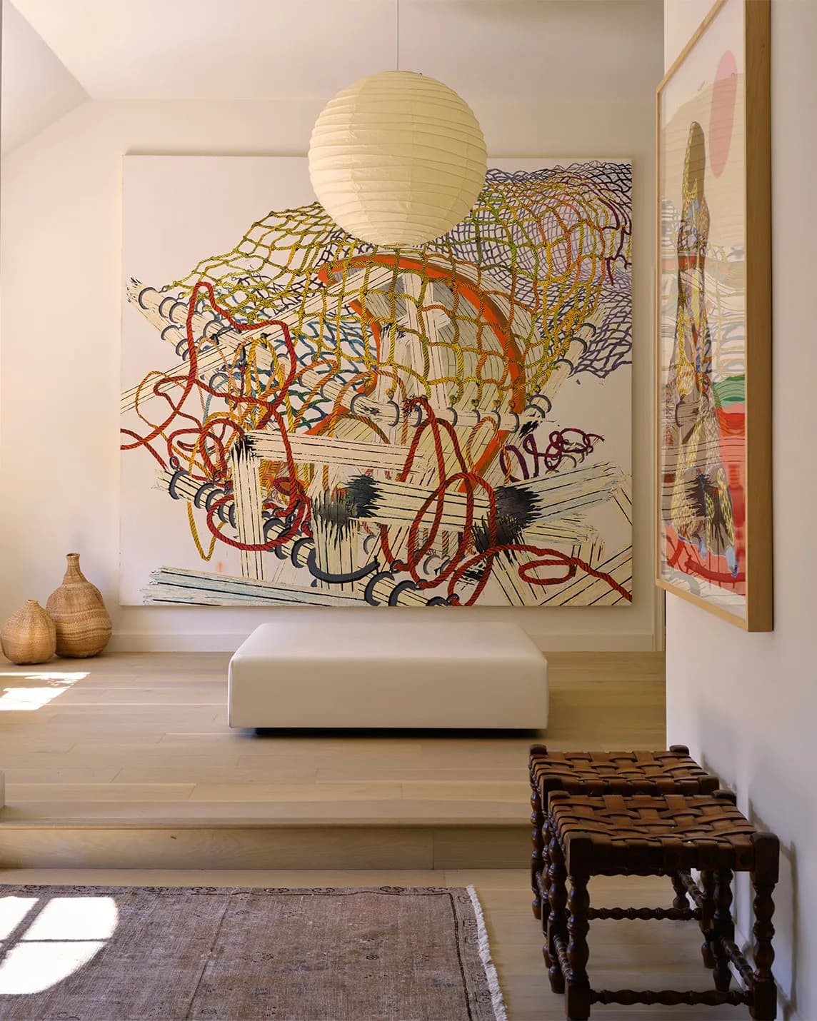 Bright contemporary room featuring a large abstract colorful painting, white round pendant light, white ottoman, woven benches, and wood floor.
