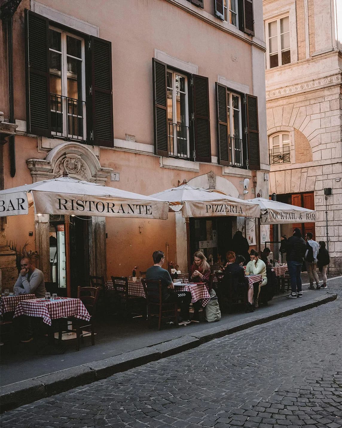 An Italian street café on a cobbled street, customers dining beneath striped awnings bearing the words ‘Ristorante’ and ‘Pizzeria’, and pink stucco buildings with shutters.