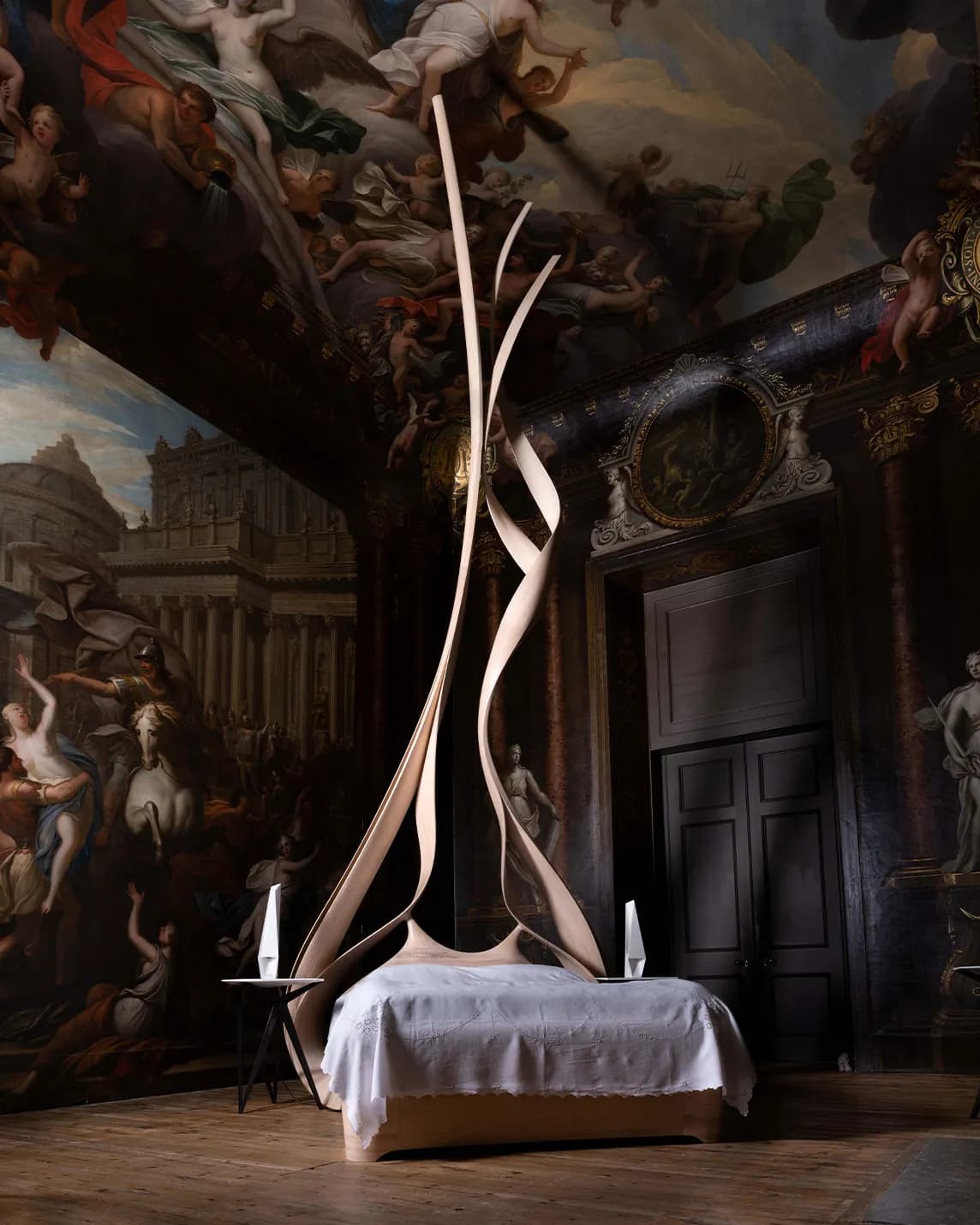 A sumptuous room with dark walls and a ceiling adorned with frescoes; a sinuous ribbon-like sculpture rises from a white bed flanked by small bedside tables.