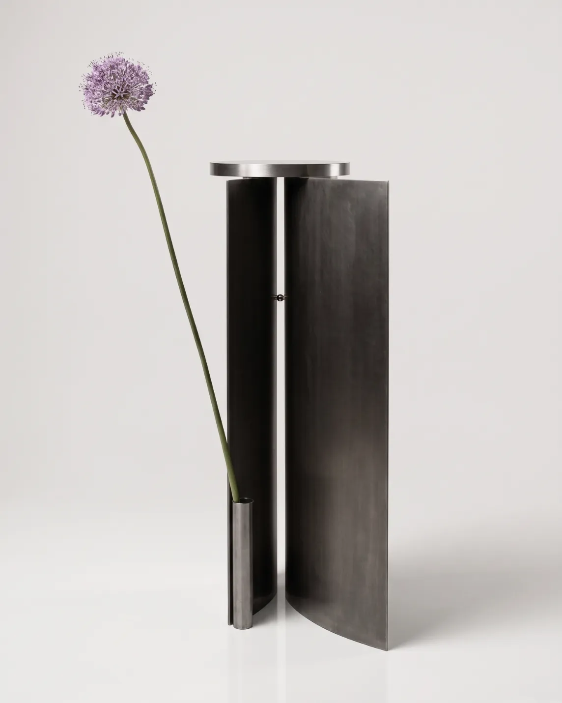 Minimalist metal sculpture with two tall dark panels and a circular top; a slim vase at the base holds a long-stemmed purple allium flower.