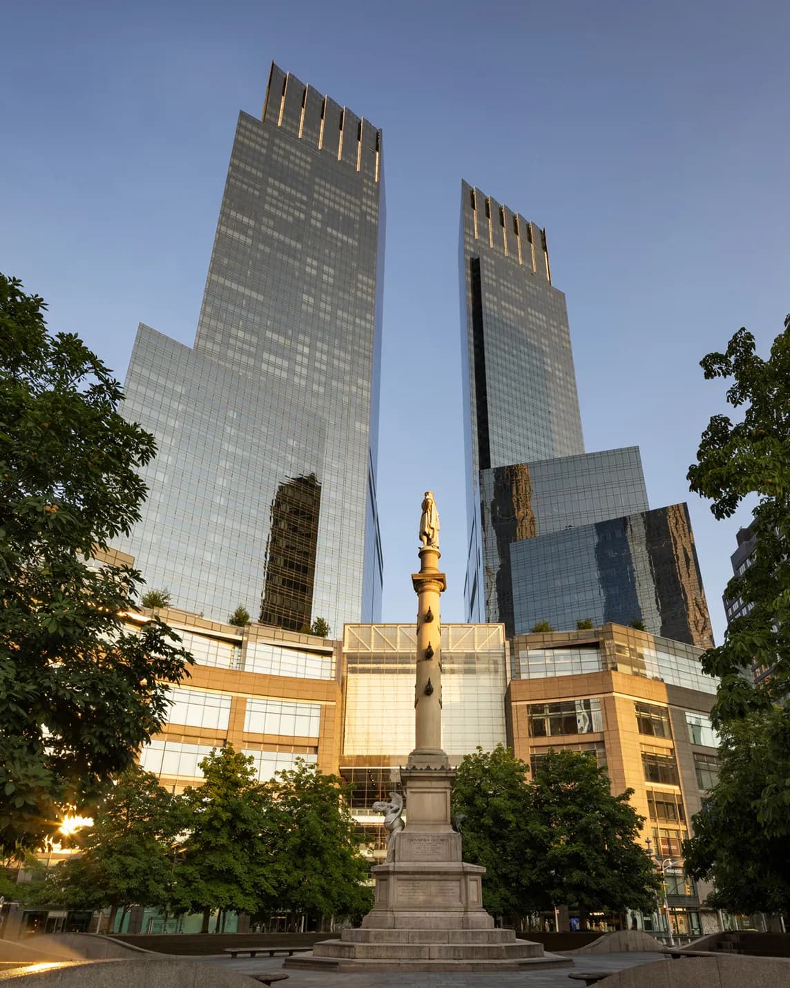 Two tall glass skyscrapers tower over a monument featuring a statue atop a column in a sunlit plaza bordered by trees.