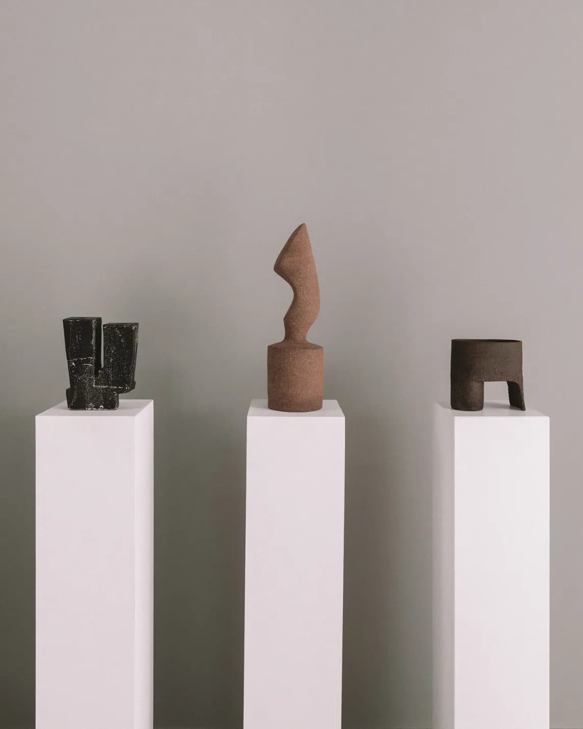 Three minimalist sculptures on white pedestals before a gray wall: left black stacked blocks, center curved brown form, right brown arch block.