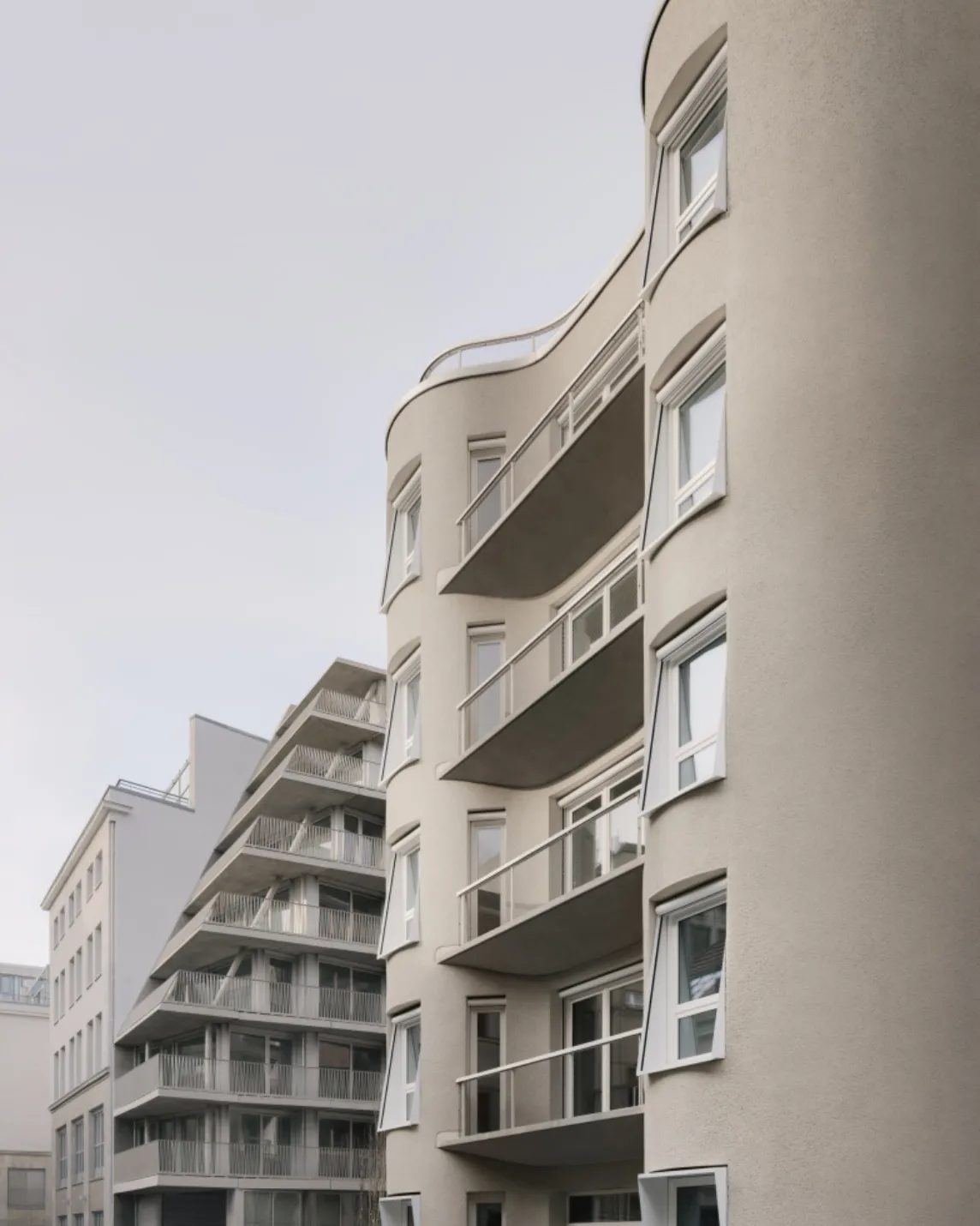 Modern beige apartment building with curved corners and protruding balconies; a gray neighboring building in the background against a pale sky.