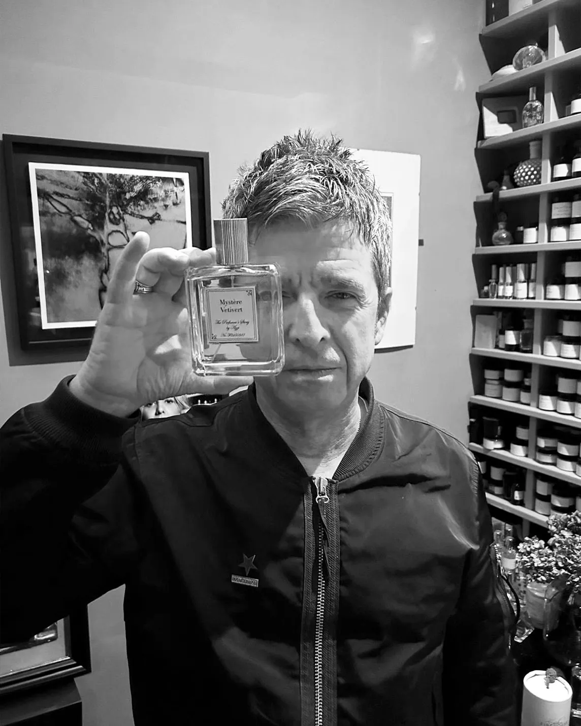 Man with short hair in a dark jacket holds a square perfume bottle up to the camera, with shelves of bottles and framed art in the background.