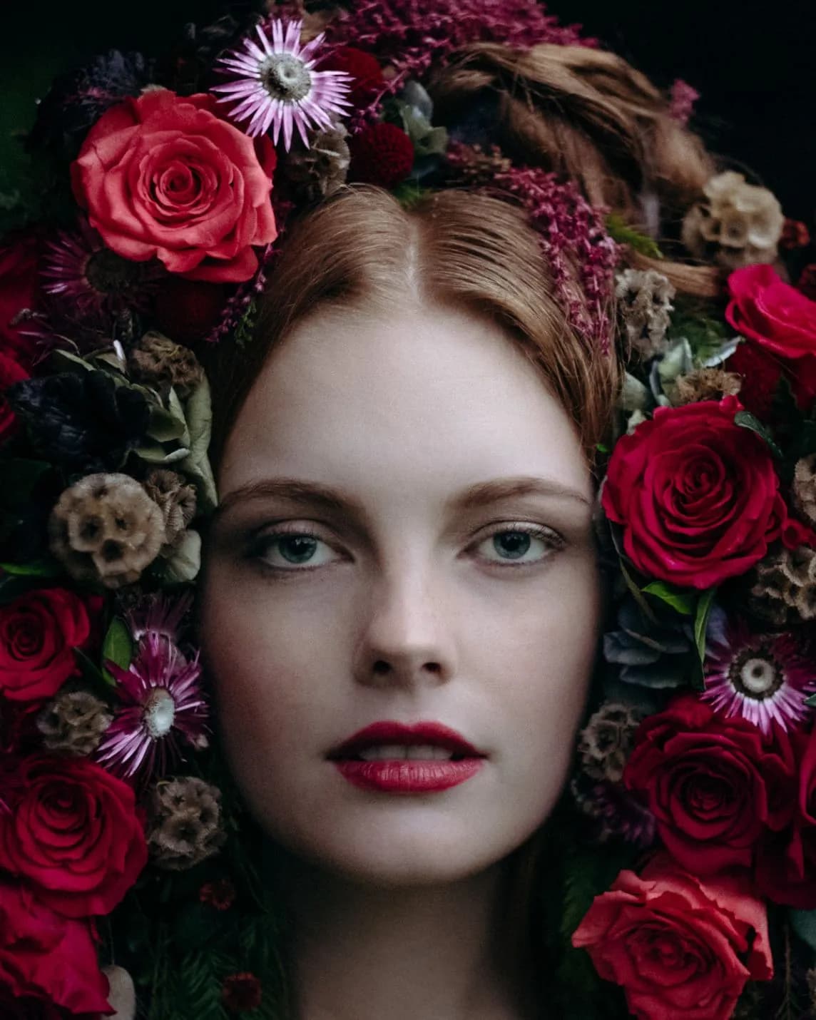 Close-up portrait of a woman with pale skin, blue eyes, and red lips, surrounded by a vibrant crown of red roses and assorted blooms.