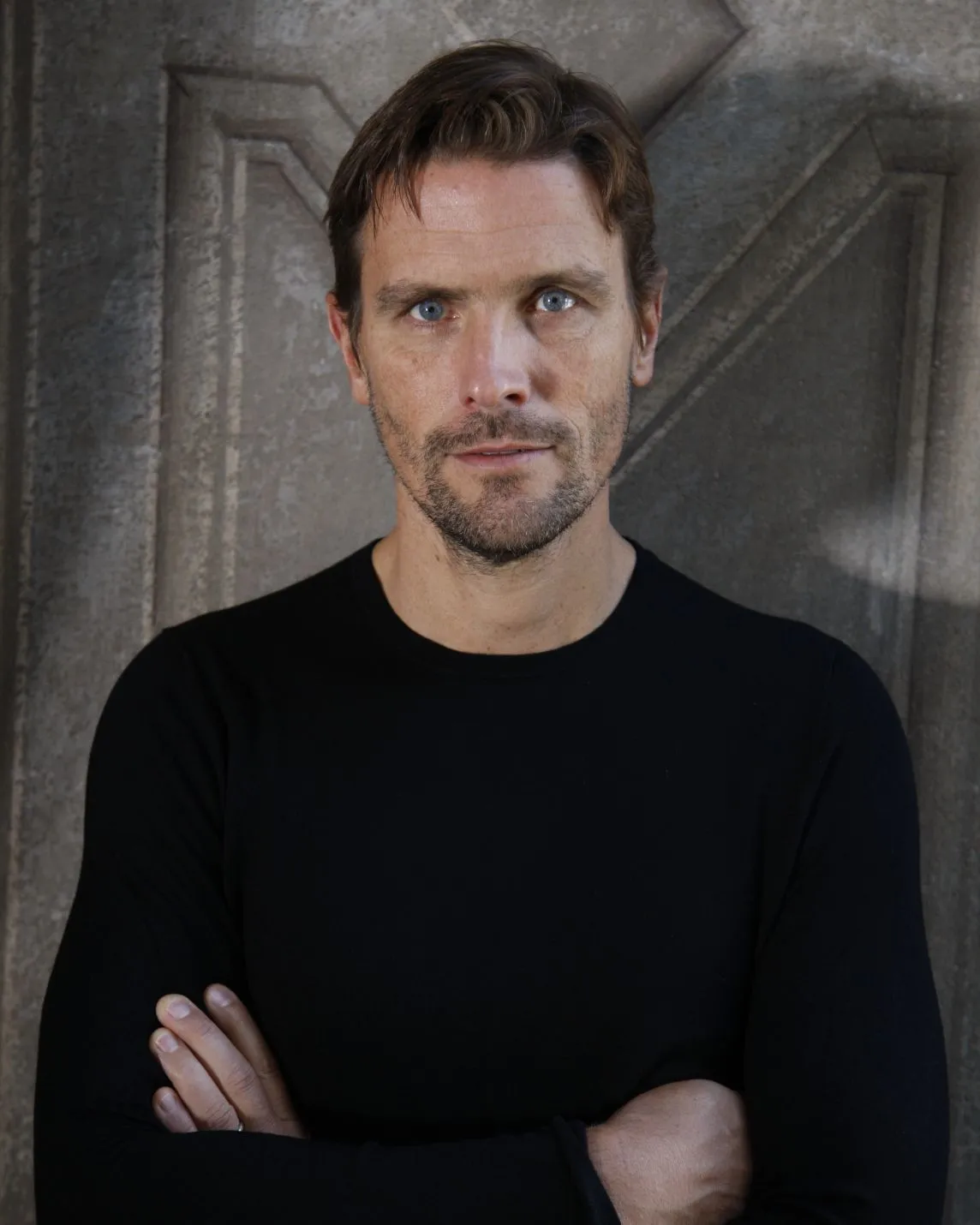 Man with short brown hair and stubble, wearing a black long-sleeve shirt, stands with arms crossed against a textured stone wall.