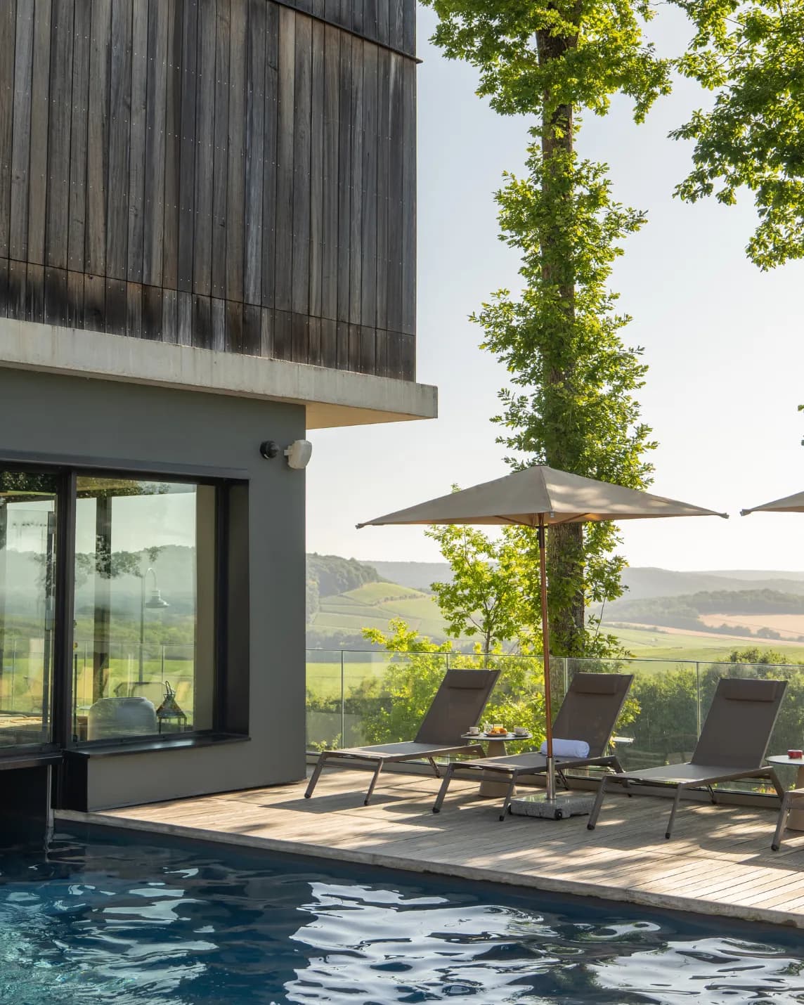 Bâtiment moderne aux boiseries sombres, jouxtant une terrasse avec piscine équipée de chaises longues et de parasols, offrant une vue sur des collines verdoyantes et des arbres.