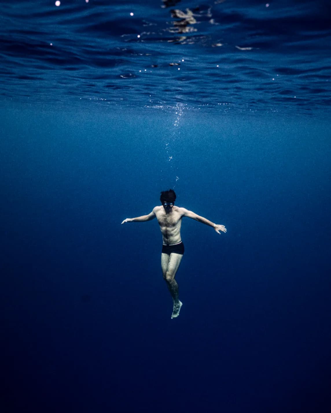 A shirtless man in dark swim trunks floats underwater with arms outstretched in the deep blue ocean.
