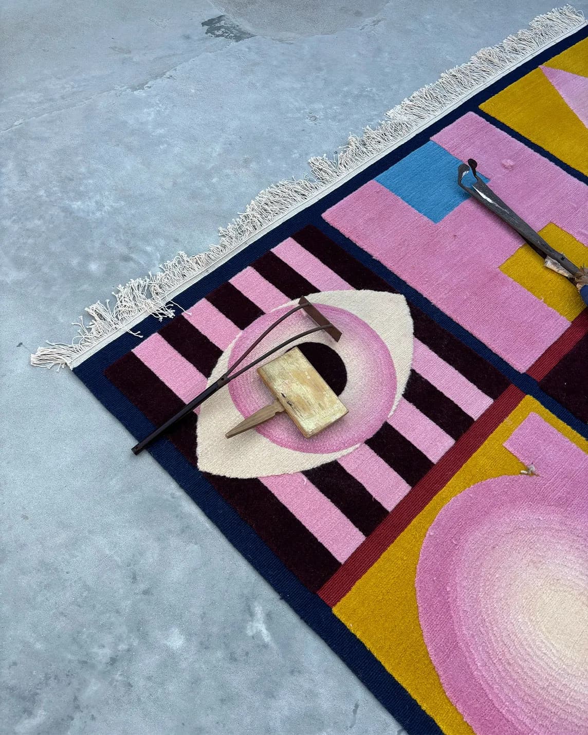 Bright geometric rug on a pale concrete floor with fringe; a pink circular object and a wooden block rest beside a curved metal tool.