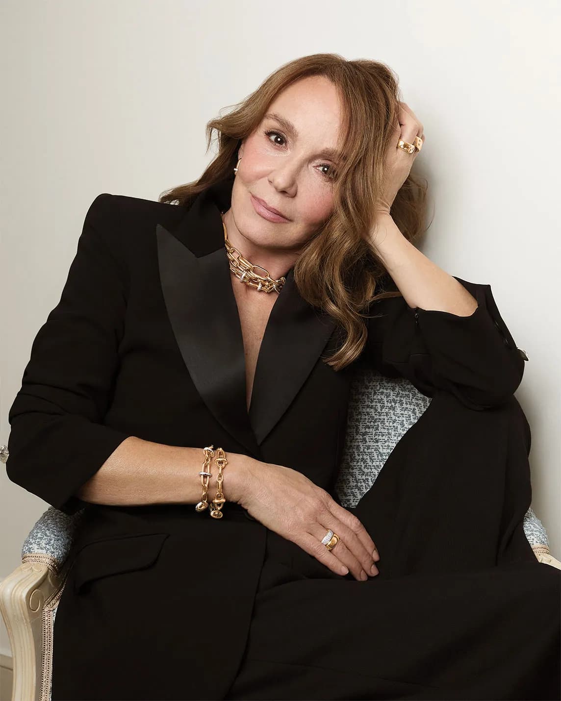 Elegant woman in a black blazer with satin lapels sits against a light background, resting her head on her hand, adorned with gold jewelry.