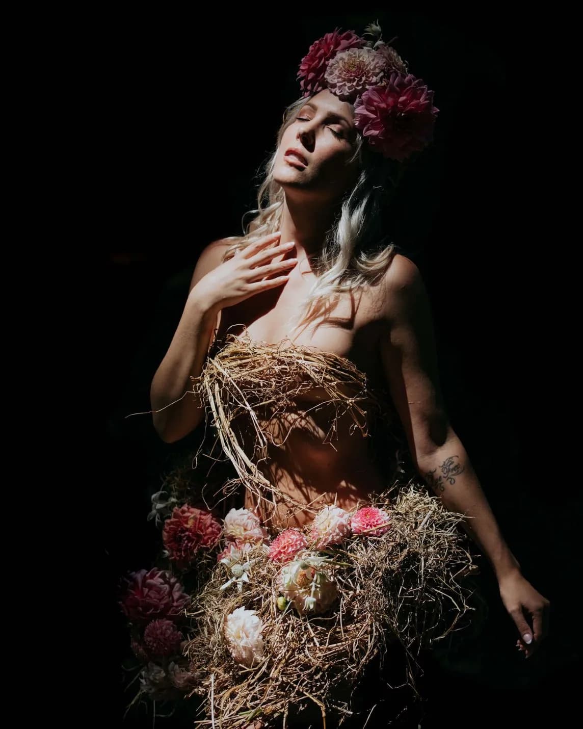 Woman with closed eyes wearing a large pink flower crown; bare shoulders and a nest-like straw-and-flower skirt against a dark background.