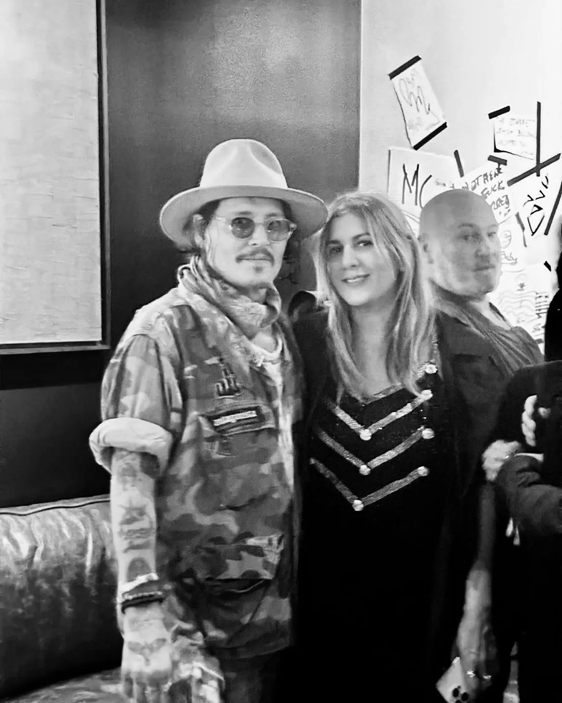 Man in wide hat, sunglasses, scarf and camo jacket poses with woman in black sequined dress; posters visible in background.