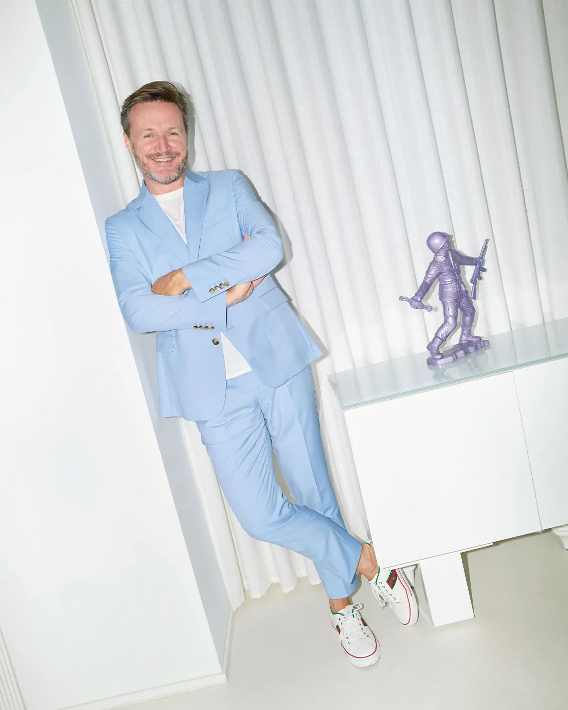 Smiling man in a light blue suit leans against a white wall with arms crossed, beside a shelf holding a purple knight statue; white sneakers.