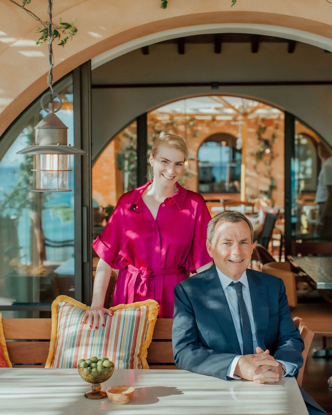 Smiling woman in a magenta dress stands beside a seated man in a suit at an outdoor cafe; olives and striped cushions sit on the table.