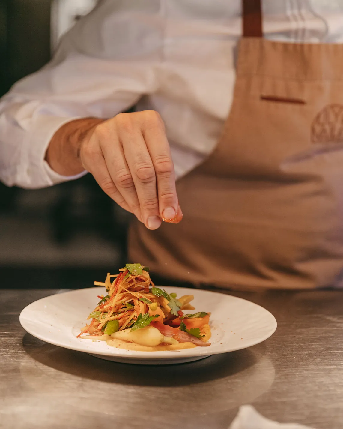 Chef sprinkling garnish over a plated dish of julienned vegetables with herbs on a white plate.