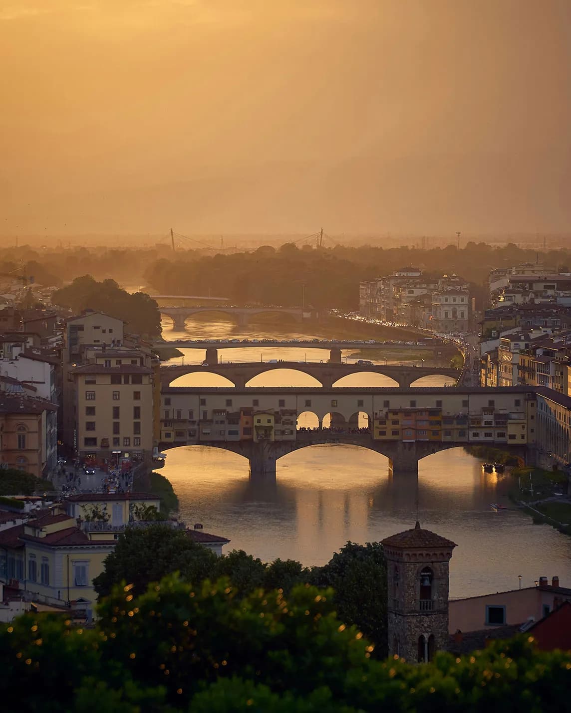 A golden sunset over a town with arched bridges spanning a tranquil river, colourful buildings along the riverbank and a tower in the foreground.