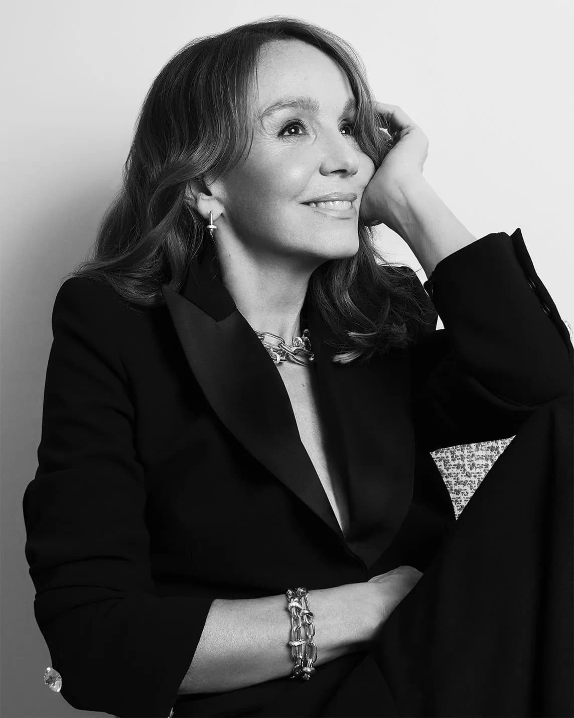 Black-and-white portrait of a smiling woman in a blazer, resting her head on her hand, wearing a chunky necklace, earrings, and bracelet.