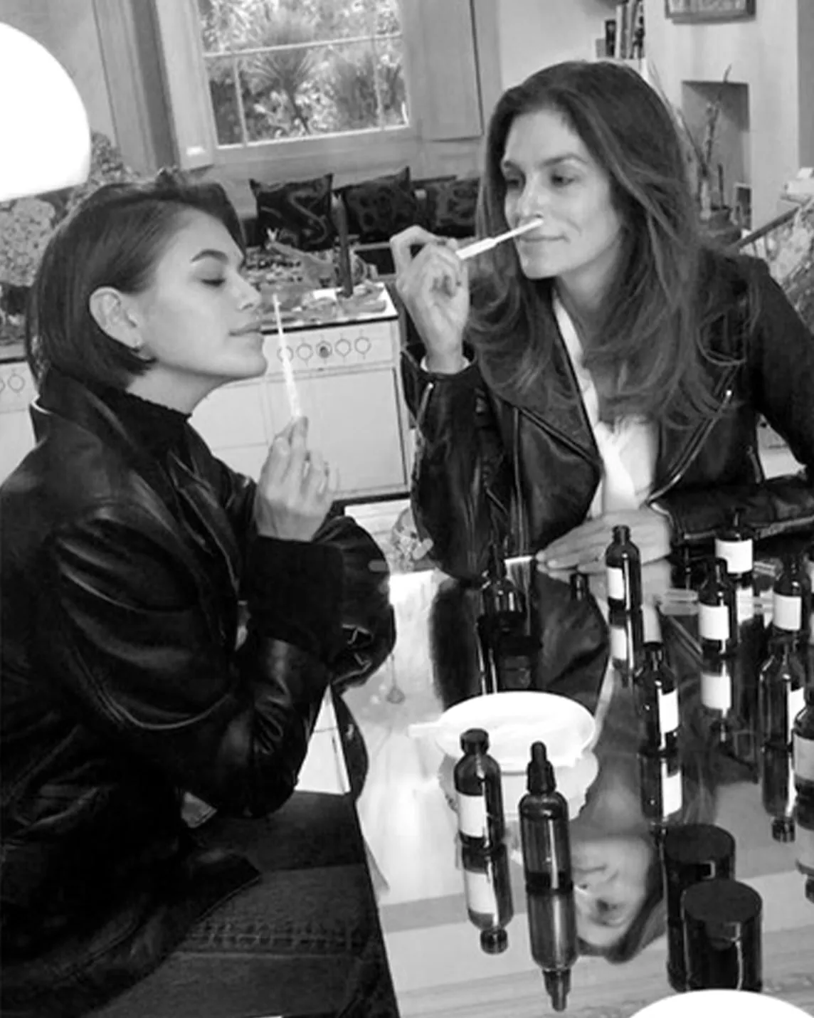 Two women in leather jackets sit at a reflective table cluttered with bottles, one applying makeup with a brush to the other’s nose.