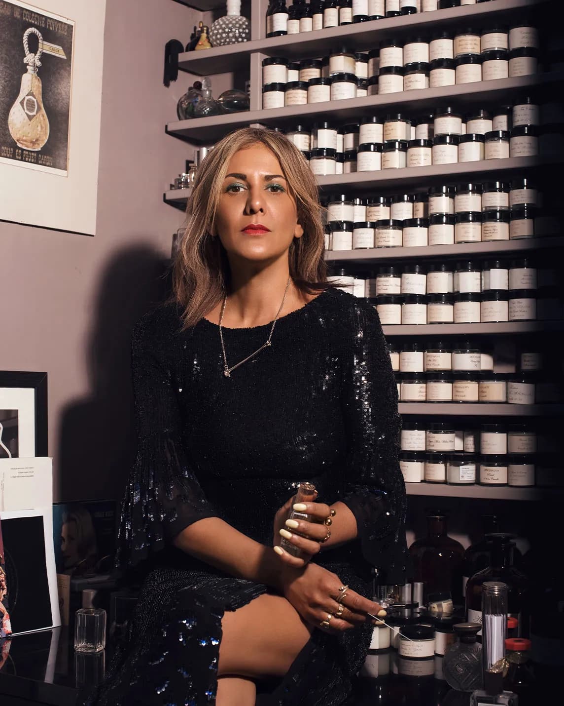 Stylish woman in a black sequined dress sits in an apothecary shop, holding a small bottle, with shelves of labeled jars behind her.