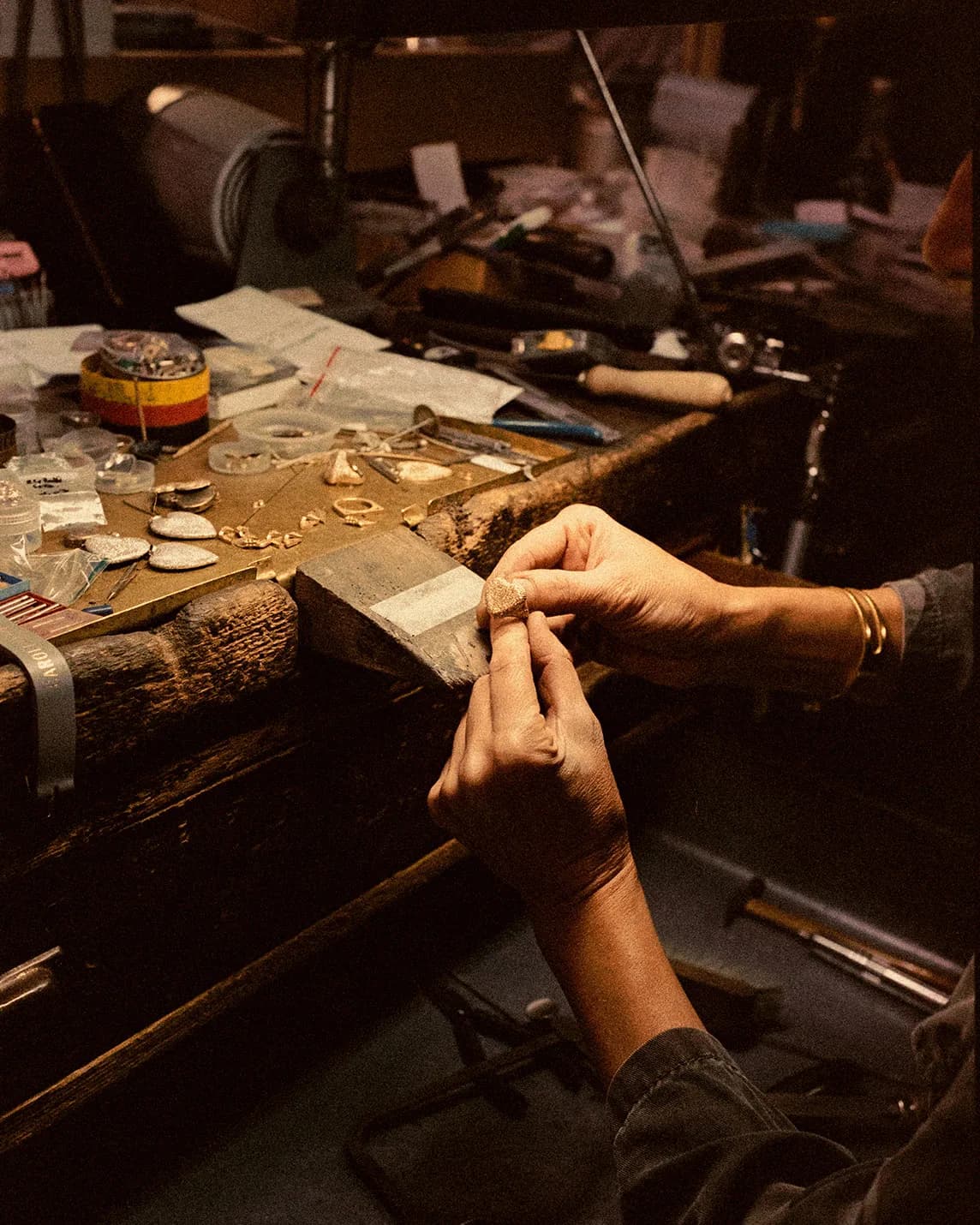 A jeweler's hands shape a small metal ring on a cluttered workbench filled with jewelry tools, stones, and components.