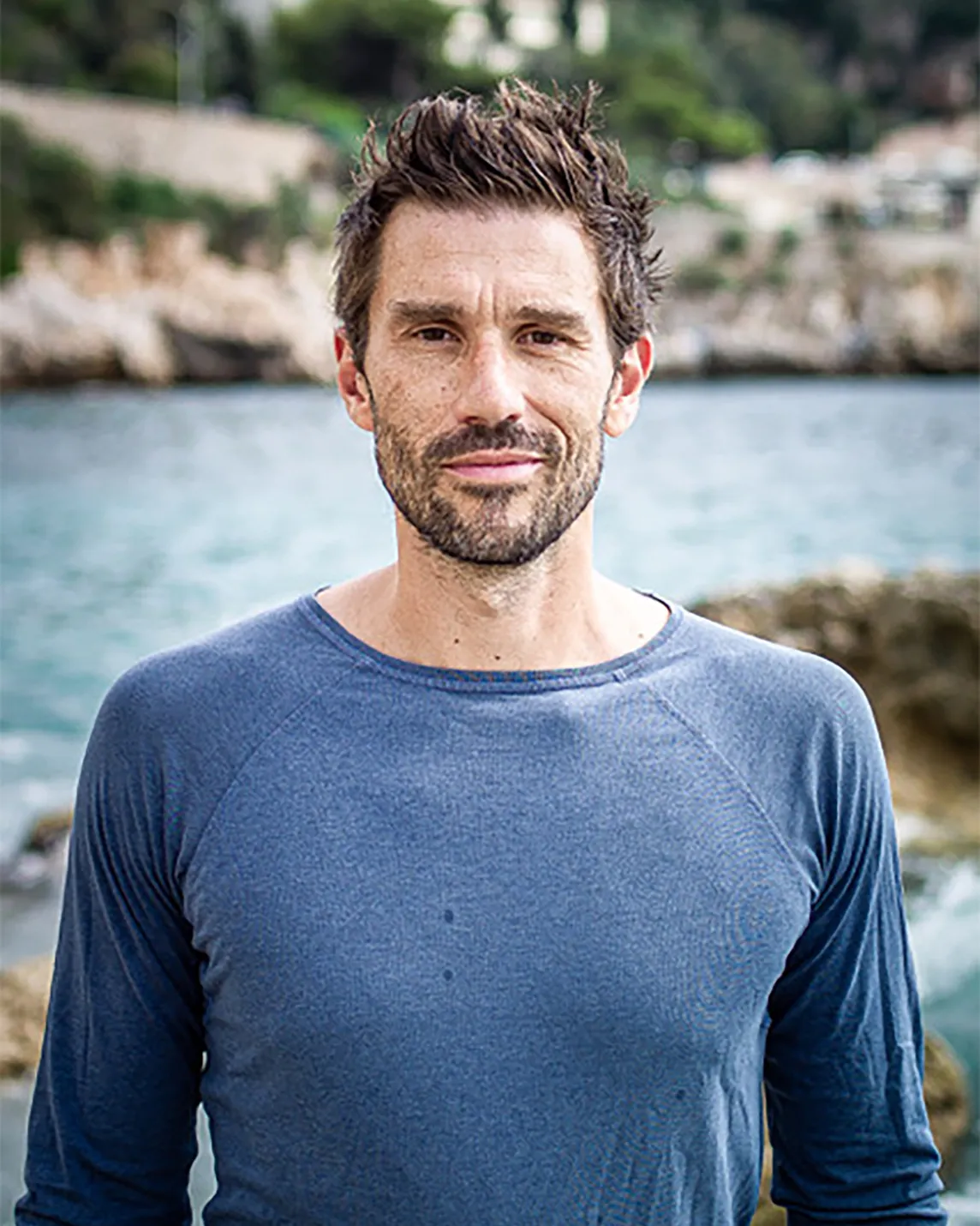 A man with short dark hair and stubble stands outdoors by a rocky coast, wearing a blue long-sleeve shirt.