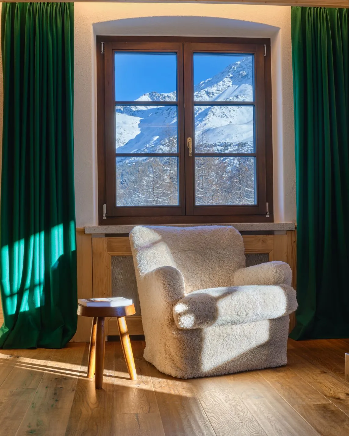 A cosy room with a window offering a view of snow-capped mountains; on the warm wooden floor stand a fluffy white armchair and a small wooden stool, along with green curtains