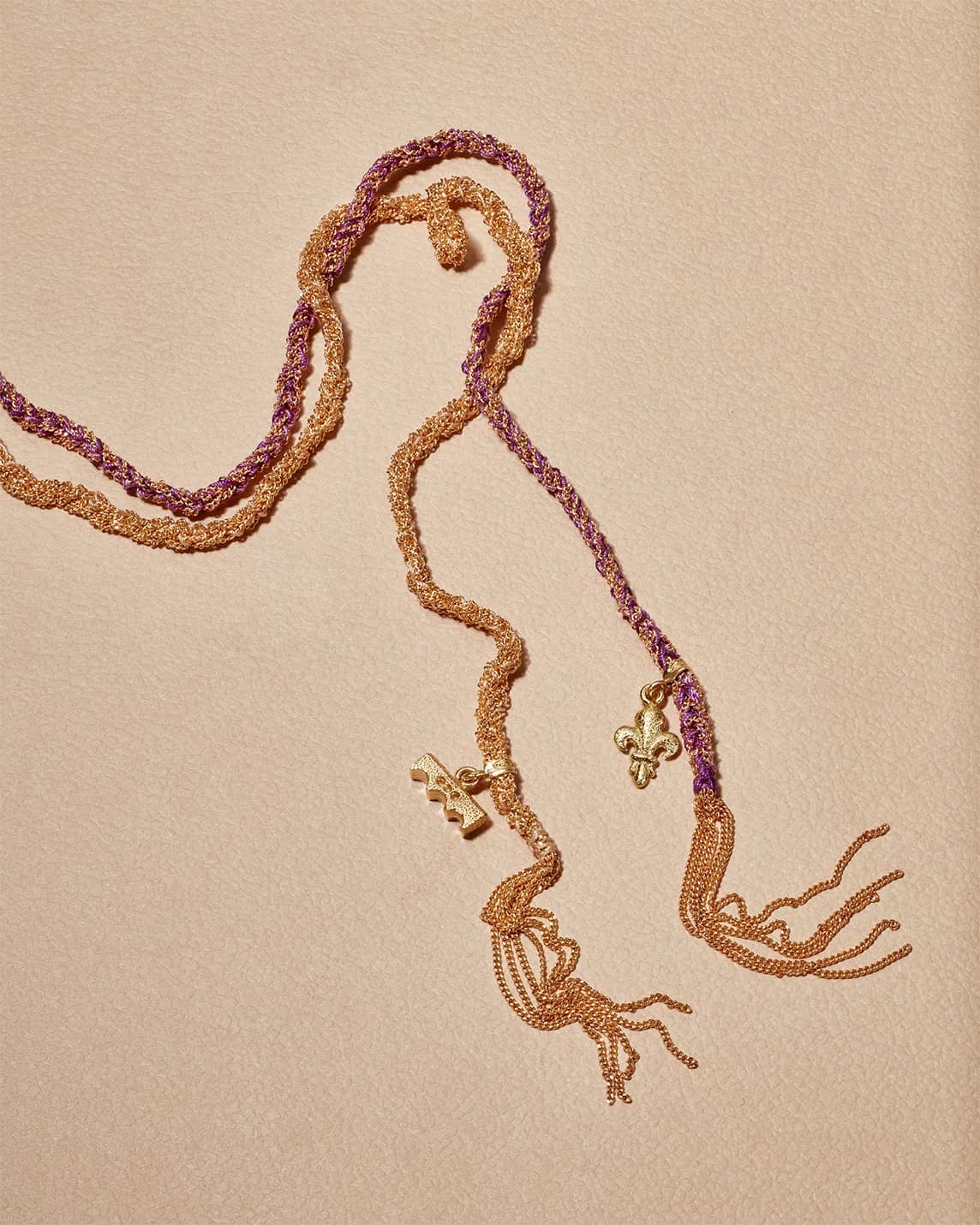 Two-tone gold and purple beaded necklace, intertwined strands with tassels and fleur-de-lis charms on a beige background.