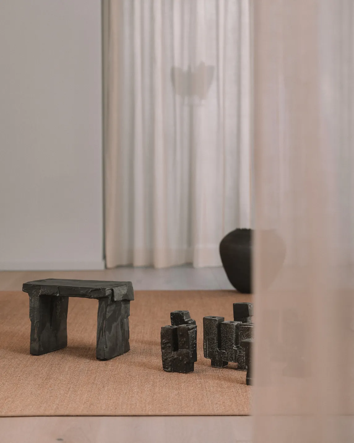 Minimalist room with a dark stone bench and several black stone blocks on a beige rug, pale curtains in the background.