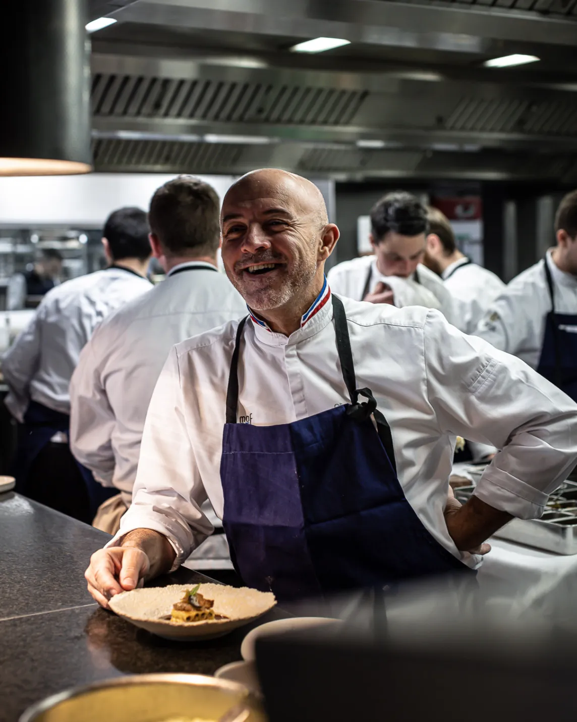 Smiling chef in a white jacket and dark apron leans at a stainless kitchen counter, presenting a plated dish as other cooks work in the background.