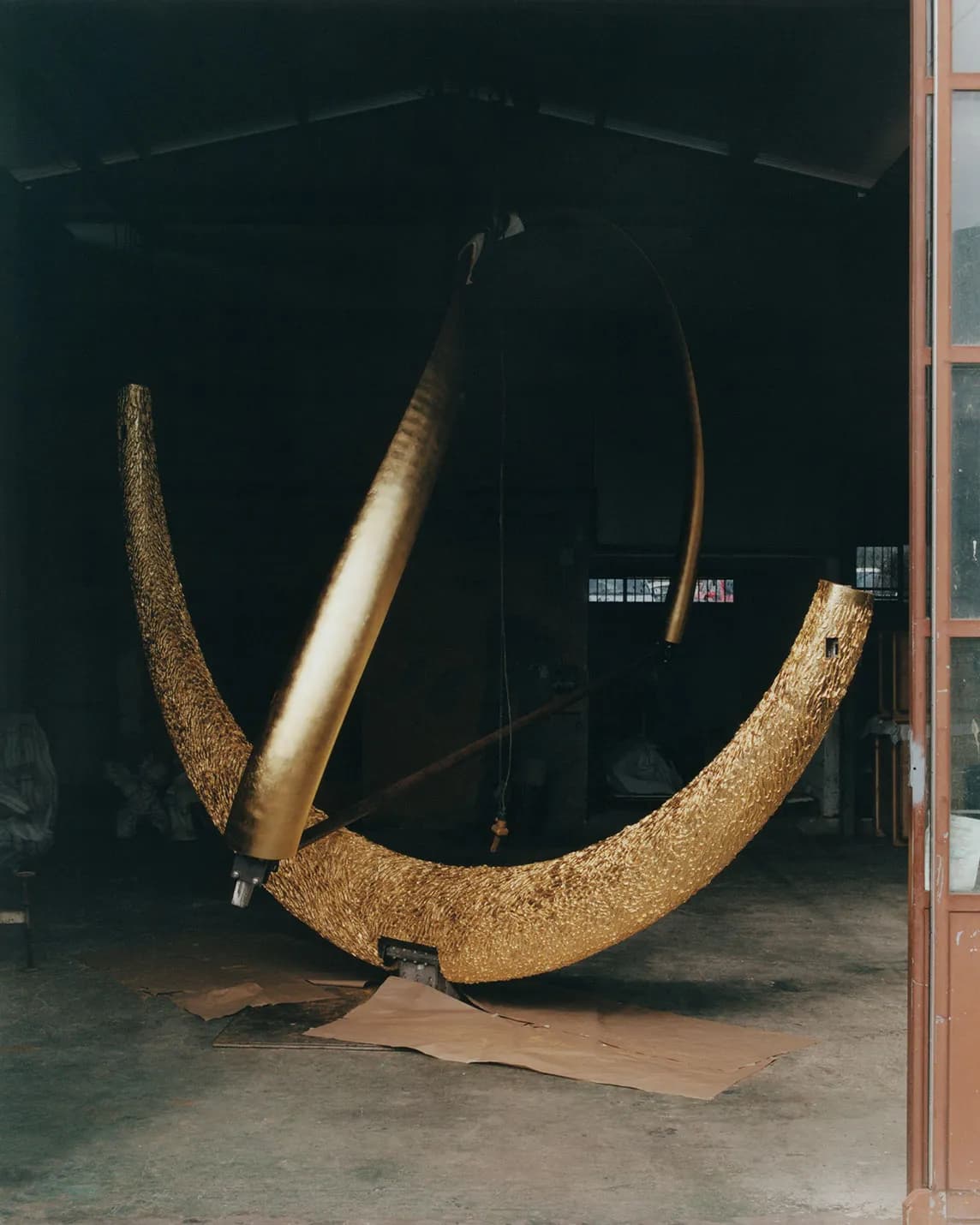 A dimly lit workshop with a large, textured, golden crescent-shaped sculpture on the floor and a smooth, curved golden rod arching over it.