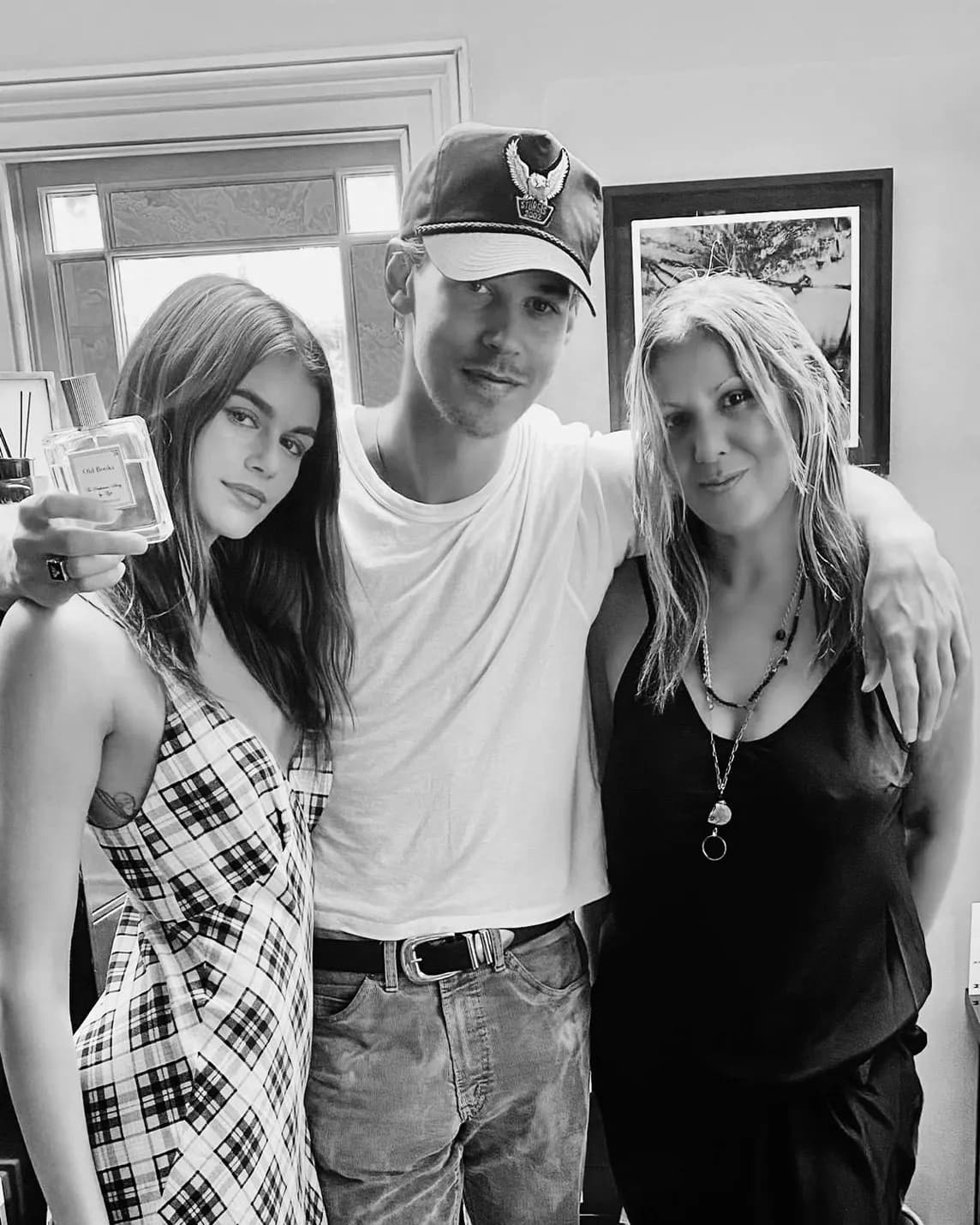 Black-and-white photo of three friends indoors: left woman in a plaid dress holding a perfume bottle, center man in a cap, right woman in a black top.