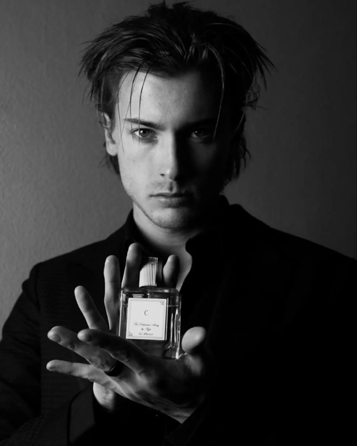 Black-and-white portrait of a man with tousled hair in a dark suit, presenting a perfume bottle toward the camera with a serious gaze.