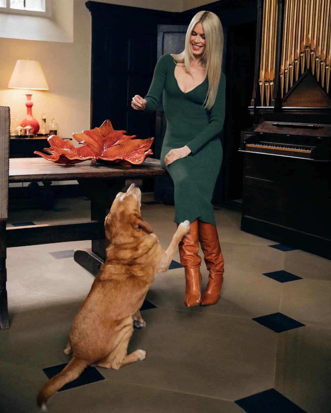 A woman in a green dress and brown knee-high boots dances with a golden retriever indoors beside a table with red leaf decor and a large organ.