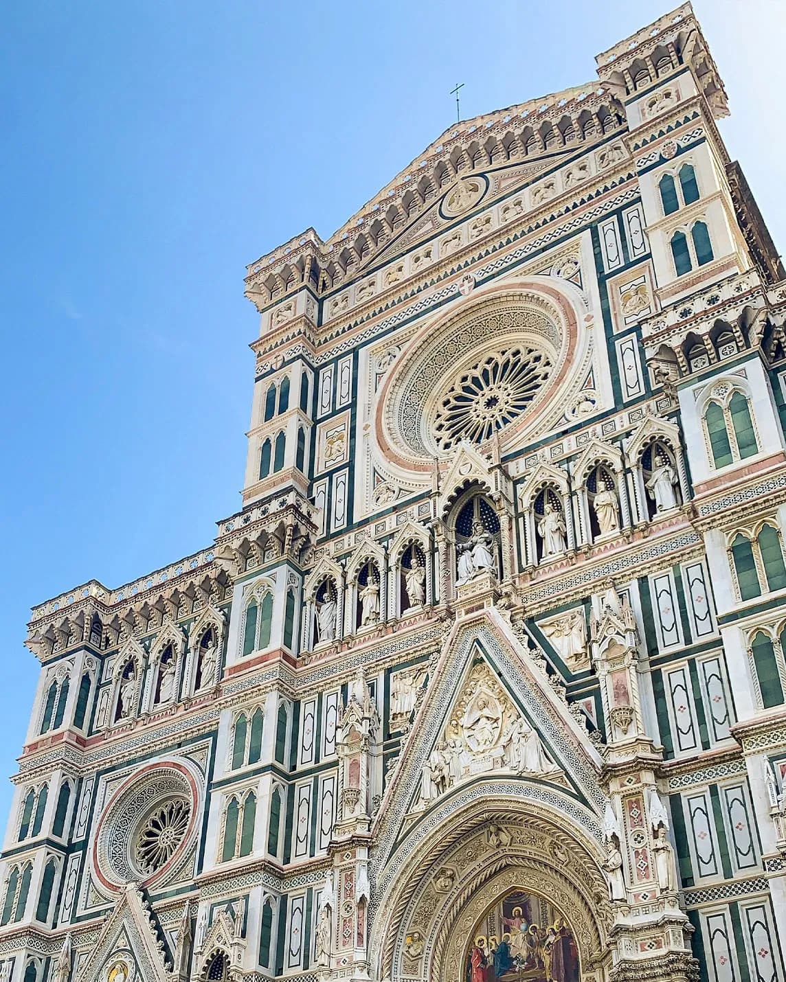 An ornate Gothic cathedral façade made of green, white and pink marble, featuring a large rose window and sculpted arches set against a blue sky.