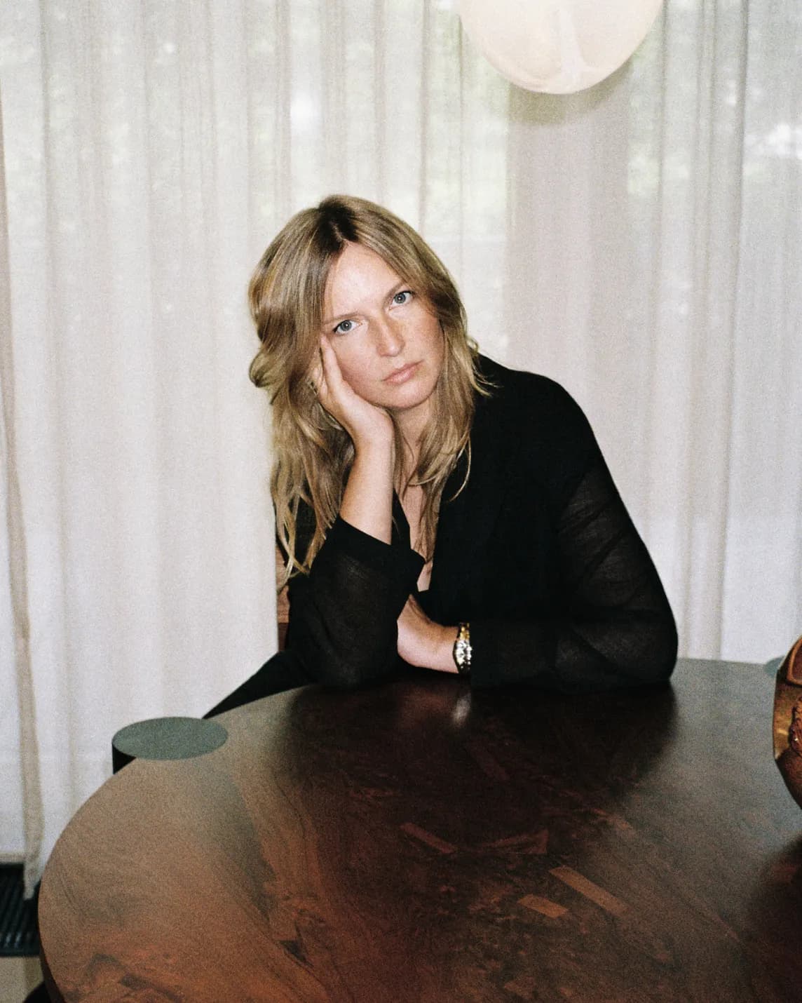 Blonde woman sits at a round wooden table indoors, head resting on her hand, wearing a black sheer top, white curtains in the background.