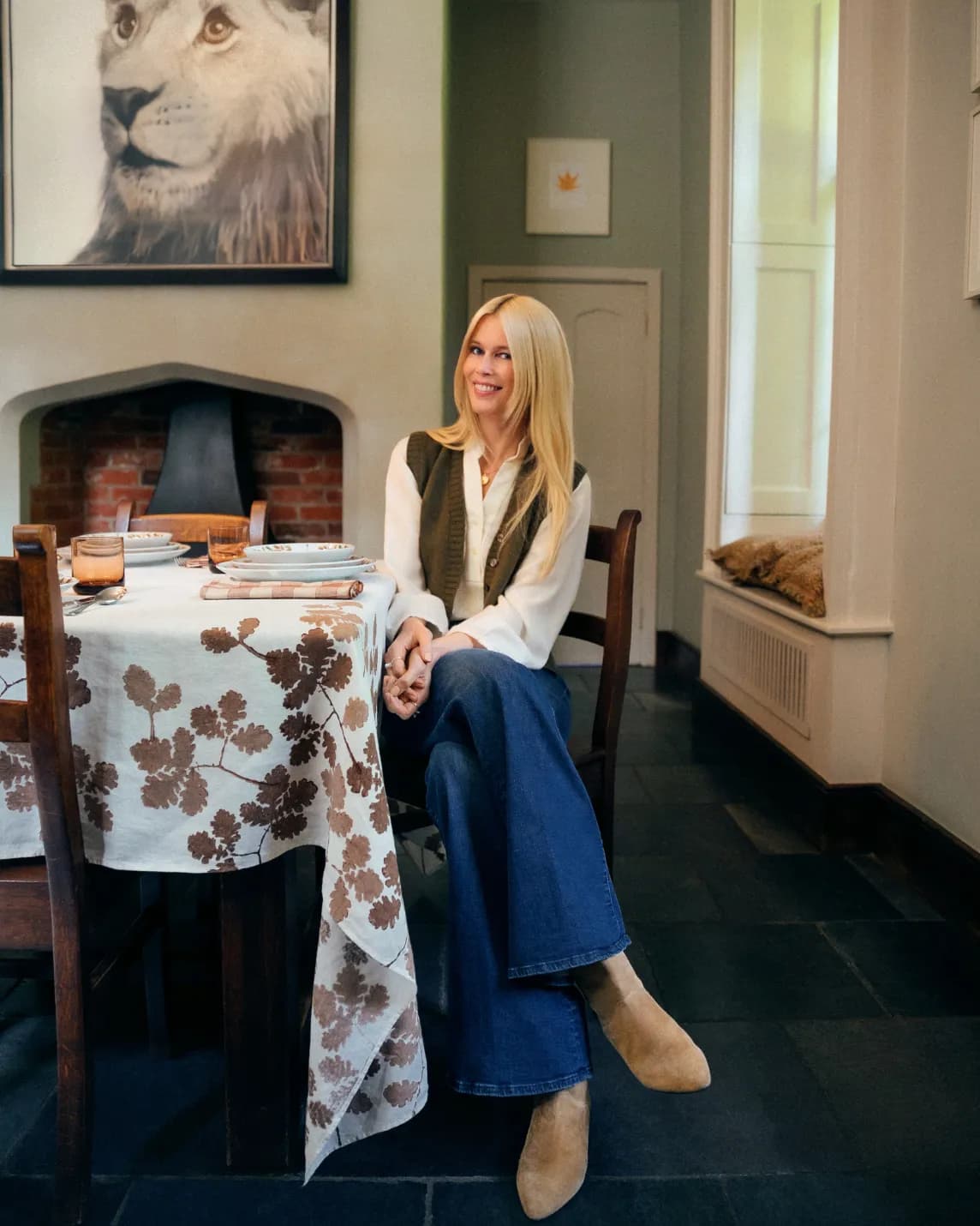 Smiling blonde woman in white blouse and vest with jeans sits at a table with a floral cloth in a cozy dining room; lion portrait above the fireplace.