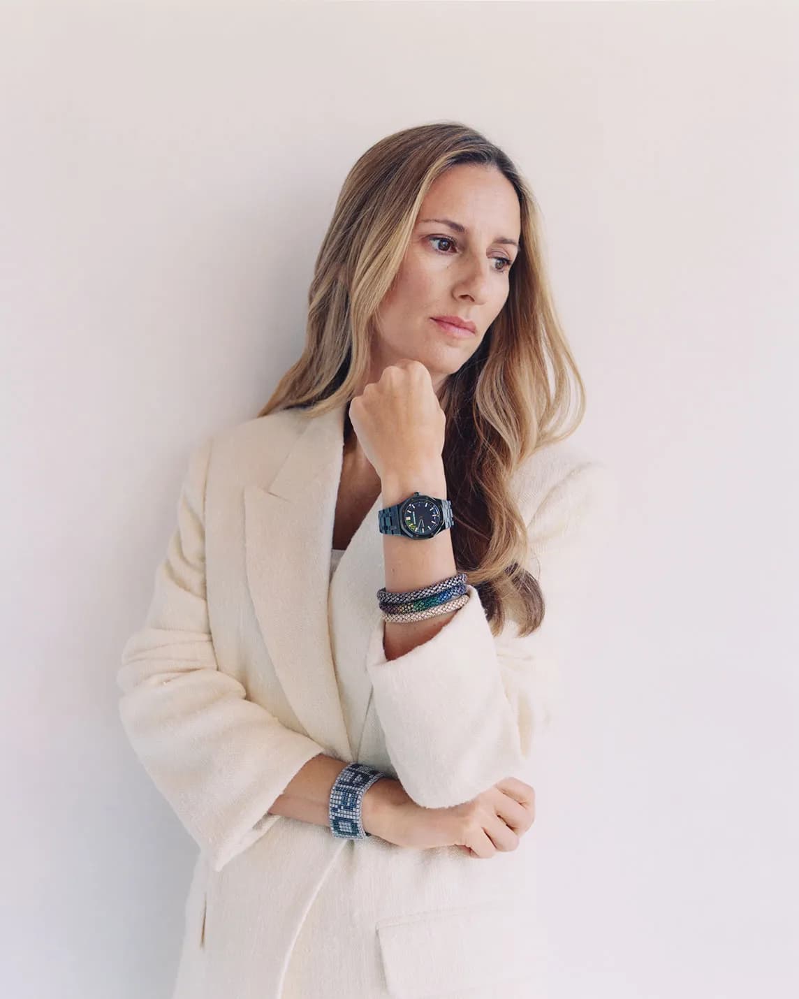 Thoughtful woman with long blonde hair in a cream blazer, resting her chin on her hand; blue watch and bracelets on her wrist.