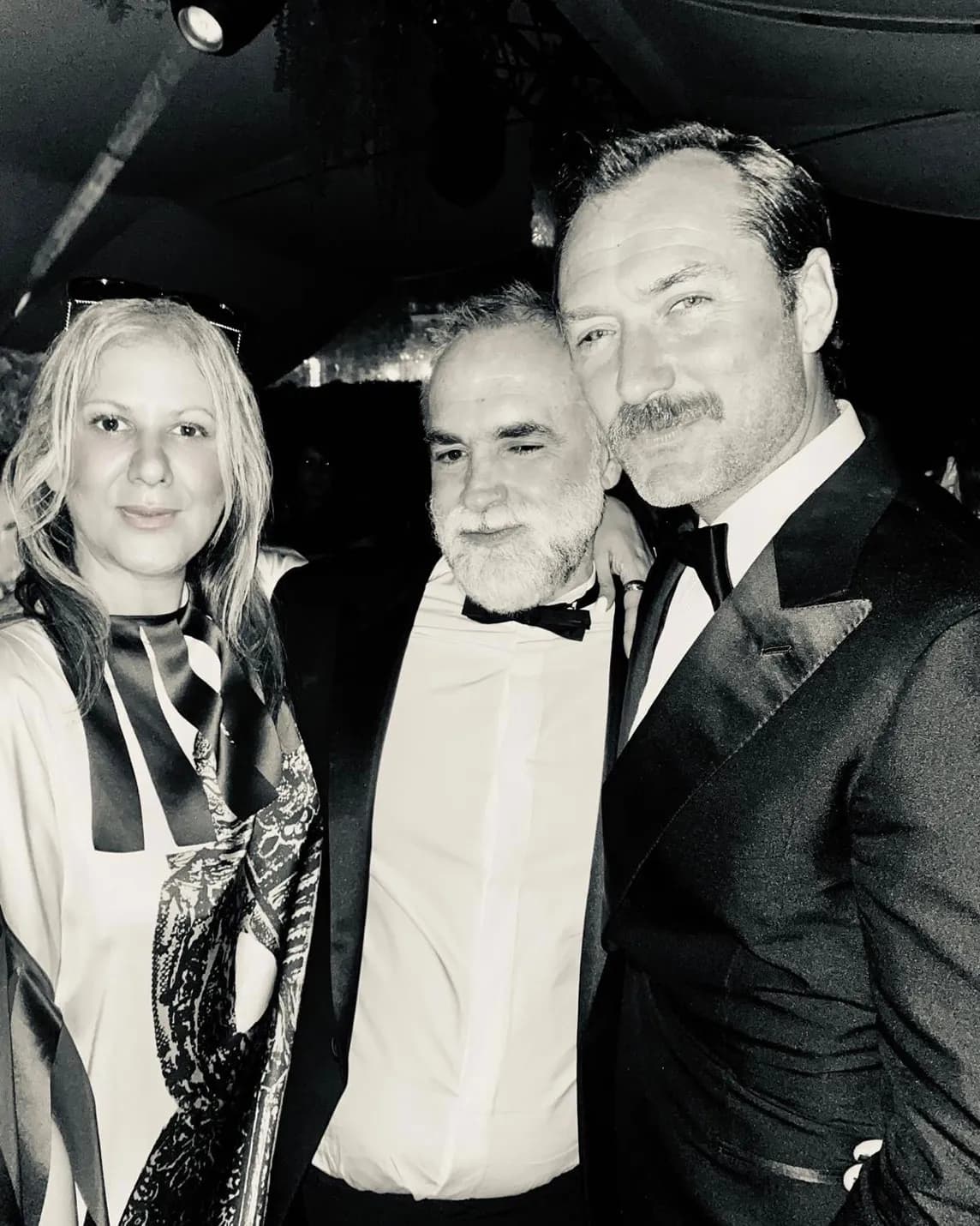 Black-and-white photo of three people in formal attire posing closely together: a woman on the left and two men in tuxedos.