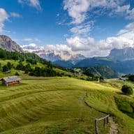 Vista panoramica di una valle verde e lussureggiante con capanne in legno, circondata da montagne e alberi sotto un cielo azzurro parzialmente nuvoloso.