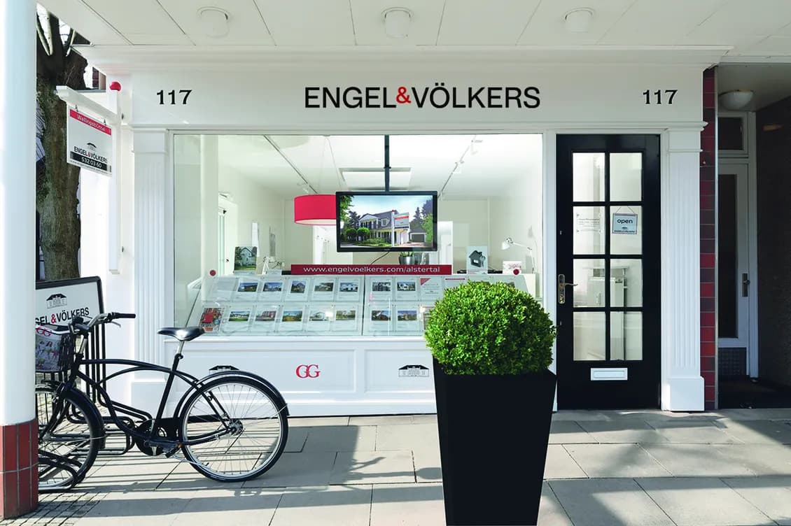 Engel & Völkers storefront with a glass brochure display, a black bicycle outside, and a tall planter with a green shrub.