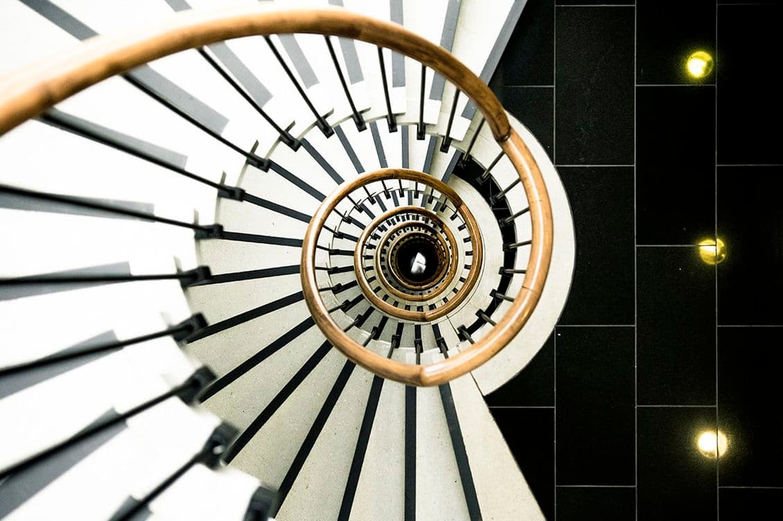 Spiral staircase with wooden handrail viewed from above, creating a circular pattern against a black and white tiled floor.