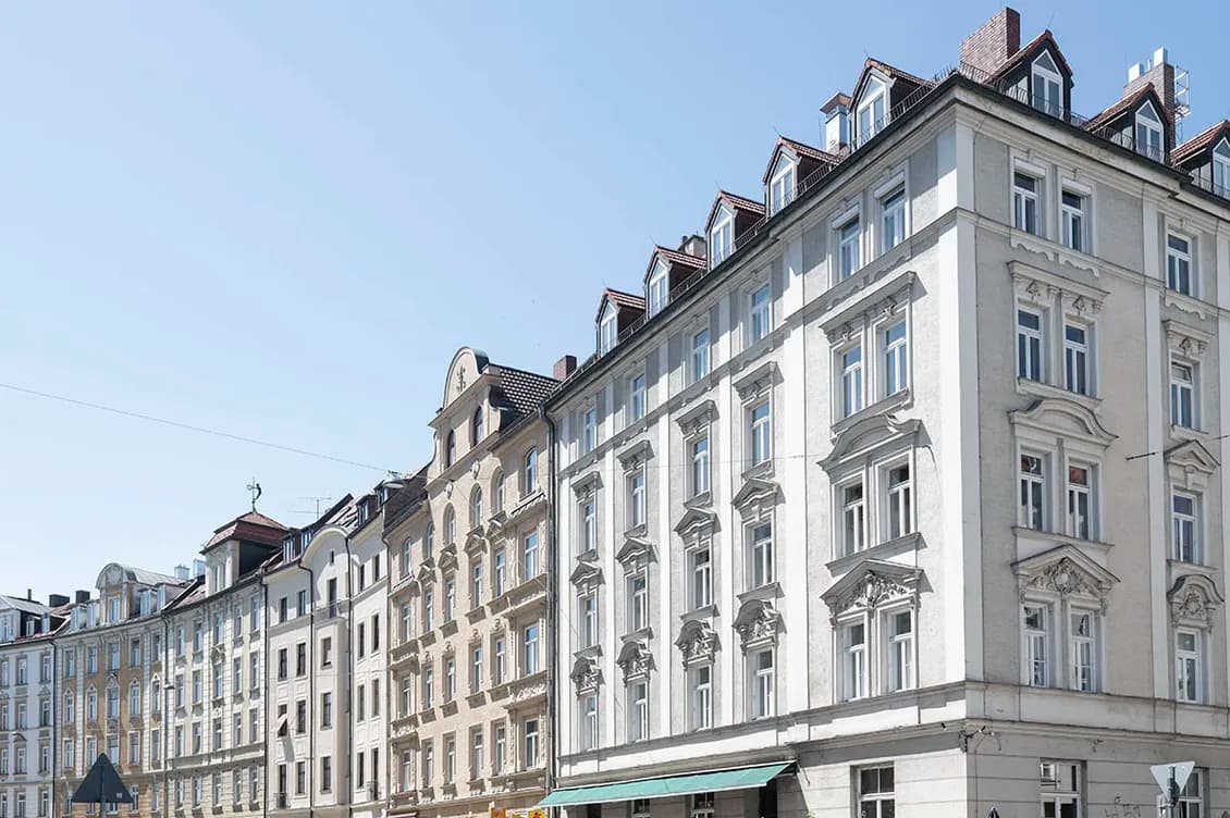 Row of elegant, historic European buildings with ornate facades under a clear blue sky. A café with outdoor seating is visible.