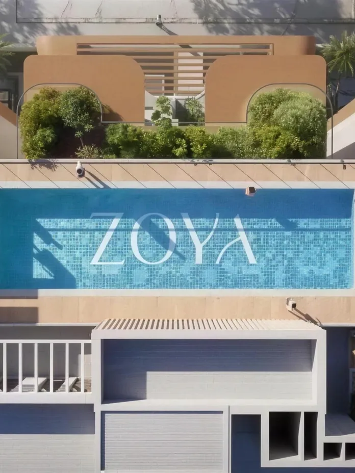 Aerial view of a rectangular rooftop pool with blue mosaic tiles forming 'ZOYA' in white letters, surrounded by tan walls, planters, and lounge areas.